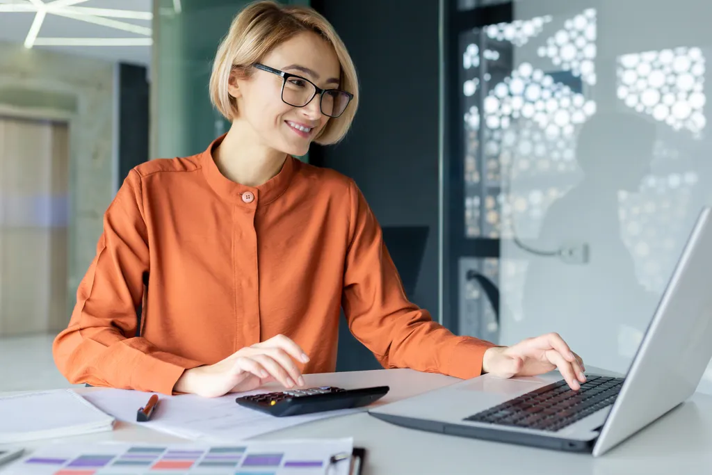 Junge Finanzierin mit Taschenrechner arbeitet im Büro am Arbeitsplatz, Geschäftsfrau hinter Papierkram zufrieden lächelnd, gute Leistungsergebnisse, Arbeit mit Verträgen, Konten und Diagrammen.
