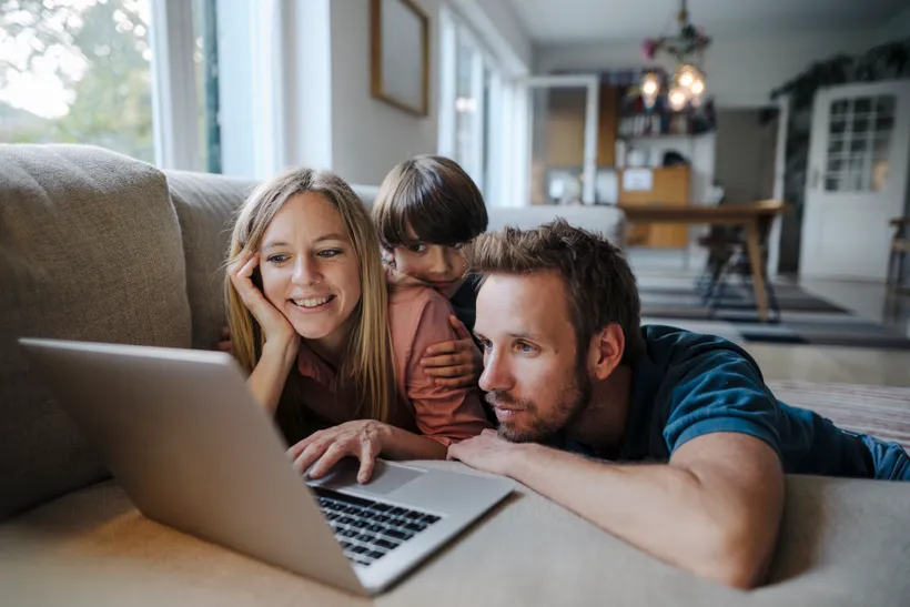 Eltern und Kind liegen auf dem Sofa im Wohnzimmer und schauen lächelnd auf einen Laptop