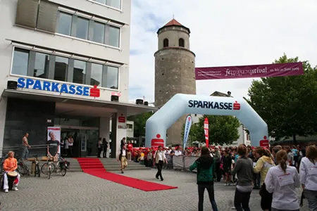 Foto der Sparkasse Feldkirch am Sparkassenplatz 1 - Menschen stehen vor Gebäude als es eröffnet wurde. Vor dem Gebäude steht ein mit aufgeblasener Bogen mit dem Wortlaut "Sparkasse" darauf